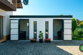 Bílý zahradní domek s pultovou střechou s černou atikou u domu