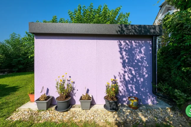 Zahradní domek s fialovou omítkou obklopený různými květinami