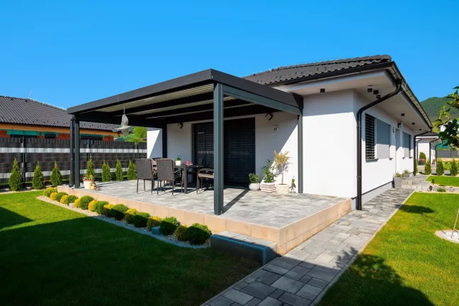 Antracitová pergola u bílého domu s dlážděnou terasou a zahradním posezením, modrá obloha