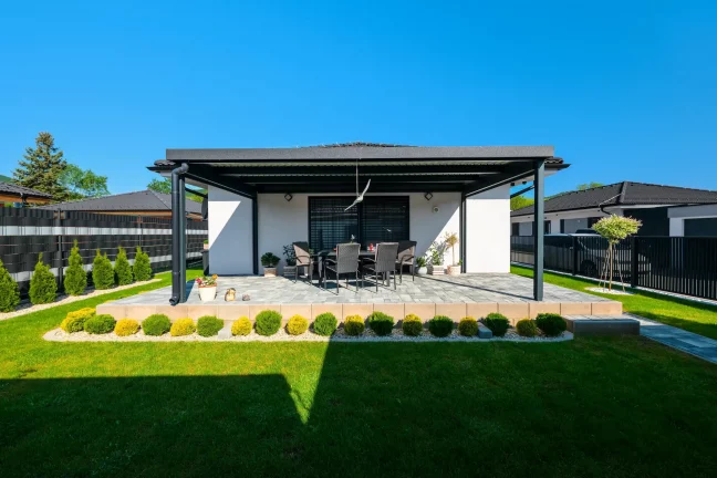Antracitová pergola s pultovou střechou, zahradní posezení a okrasné keře kolem
