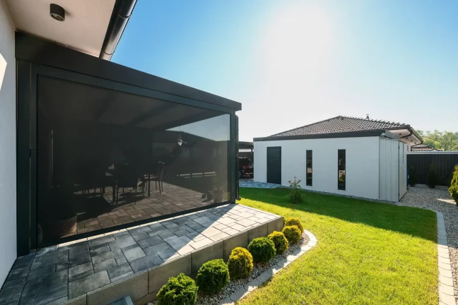 Antracitová pergola se staženou roletou u bílého domu, dlážděná terasa a zelený trávník