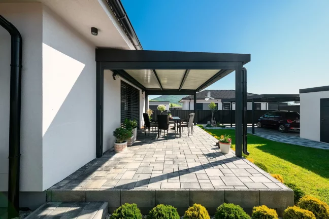 Antracitová pergola s lamelovou střechou u bílého domu, dlážděná terasa se zahradním posezením