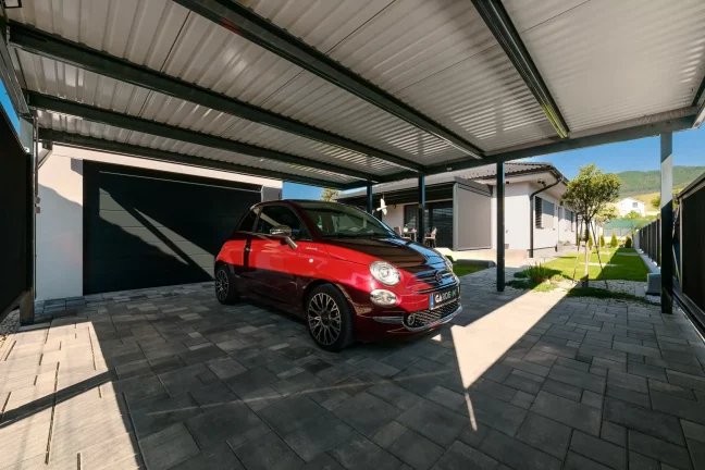 Červené auto pod přístřeškem s pultovou střechou, pohled z boku, dlážděná plocha a zelená zahrada