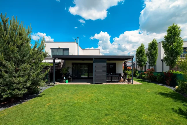 Antracitová pergola s plochou střechou u bílého domu, rozlehlý zelený trávník a stromy