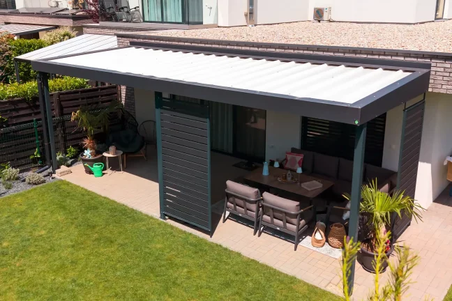 Pergola se zahradním posezením u tmavé fasády, dlážděná terasa