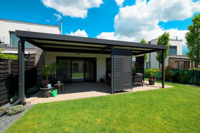 Volně stojící antracitová pergola s plochou střechou na zelené zahradě, modrá obloha s oblaky
