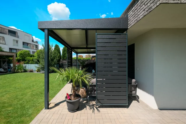 Antracitová pergola s bočním obkladem u cihlového domu, dekorativní rostliny na terase