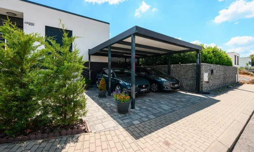 Dvě auta parkující pod antracitovým ocelovým přístřeškem s plochou střechou u domu.