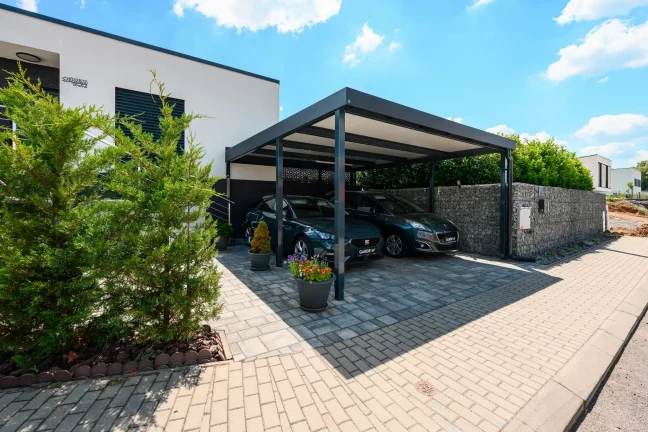 Dvě auta parkující pod antracitovým ocelovým přístřeškem s plochou střechou u domu.