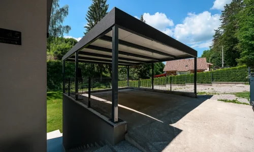 Tmavošedý přístřešek s plochou střechou ukončenou atikou u rodinného domu.