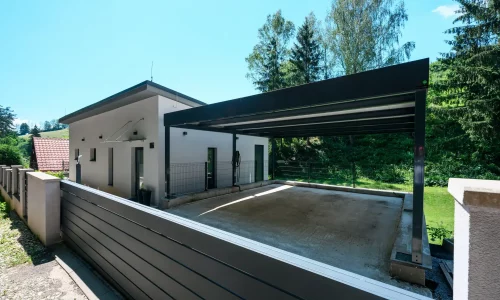 Otevřený přístřešek s antracitovou plochou střechou u bílého rodinného domu.