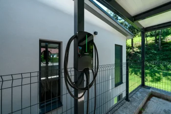 Nabíječka elektromobilů na sloupu antracitového přístřešku u omítané fasády