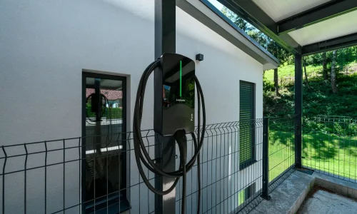 Nabíječka elektromobilů na sloupu antracitového přístřešku u omítané fasády.