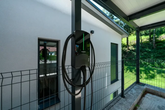 Nabíječka elektromobilů na sloupu antracitového přístřešku u omítané fasády