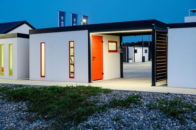 Osvětlený zahradní domek s bílou omítkou a oranžovými dveřmi Hörmann.