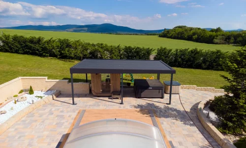 Antracitová pergola se zahradním posezením u bazénu s průhledným zastřešením, zelené kopce v pozadí