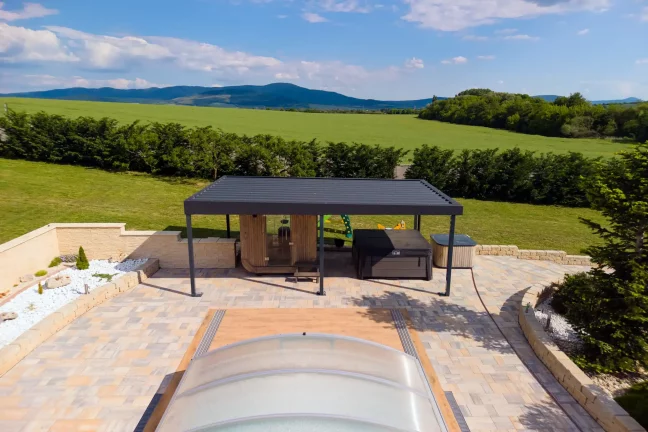 Antracitová pergola se zahradním posezením u bazénu s průhledným zastřešením, zelené kopce v pozadí