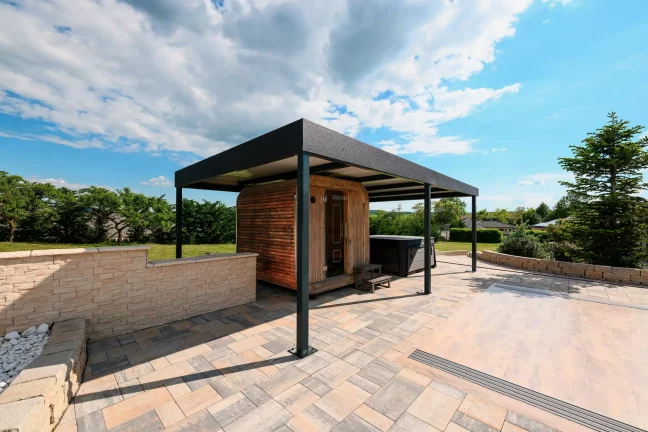Volně stojící antracitová pergola s plochou střechou na dlážděné terase, zelená zahrada v pozadí
