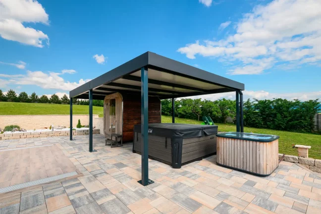 Antracitová pergola s vířivkou pod ní na dlážděné terase, zelená zahrada a modrá obloha