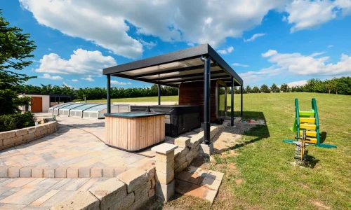 Antracitová pergola s vířivkou a dětským hřištěm v pozadí, dlážděná terasa u zahrady