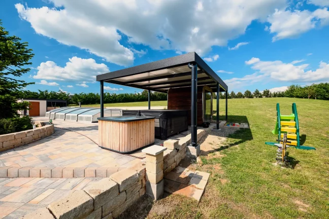 Antracitová pergola s vířivkou a dětským hřištěm v pozadí, dlážděná terasa u zahrady