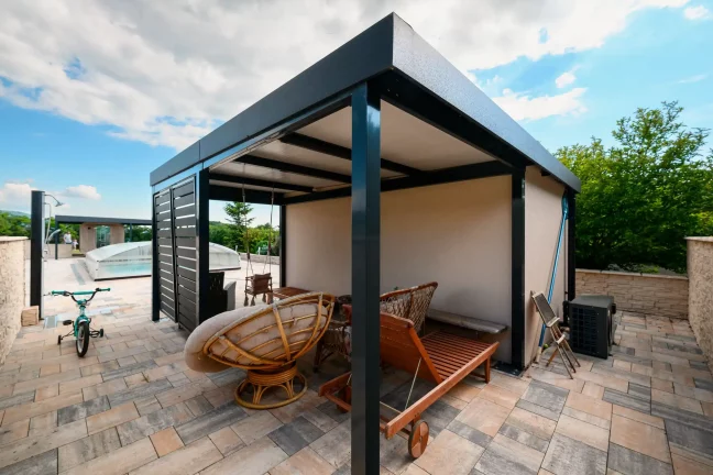 Antracitová pergola u zahradního domku s dveřmi, jízdní kolo a zahradní nábytek pod ní
