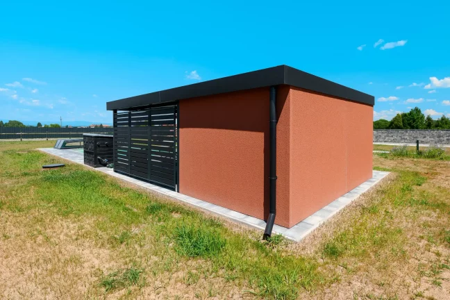 Omítka KABE cihlové barvy pokrývá stěny zahradního domku s plochou střechou.