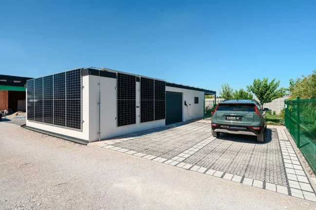 Pohled na dům se solární stěnou, zeleným autem a vraty