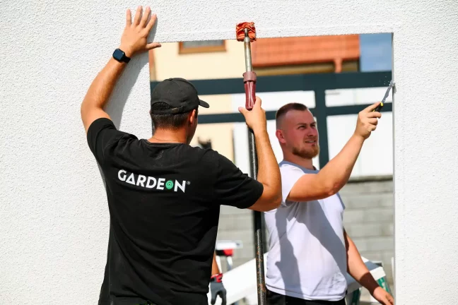 Pracovníci přitlačují velký stěnový panel a utěsňují spáru silikonem