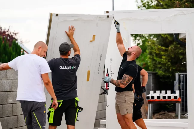 Tři pracovníci zvedají velký očíslovaný stěnový panel ke konstrukci