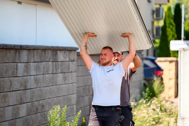 Dva pracovníci nesou nad hlavami velký krémový střešní panel