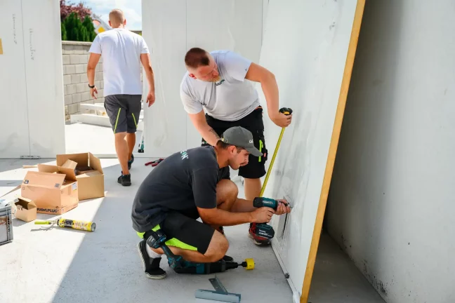 Tři montážníci měří a vrtají bílý stěnový panel na zemi